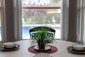 A dining table with a potted plant in the center, two bowls on either side, and a view of a swimming pool through the window.