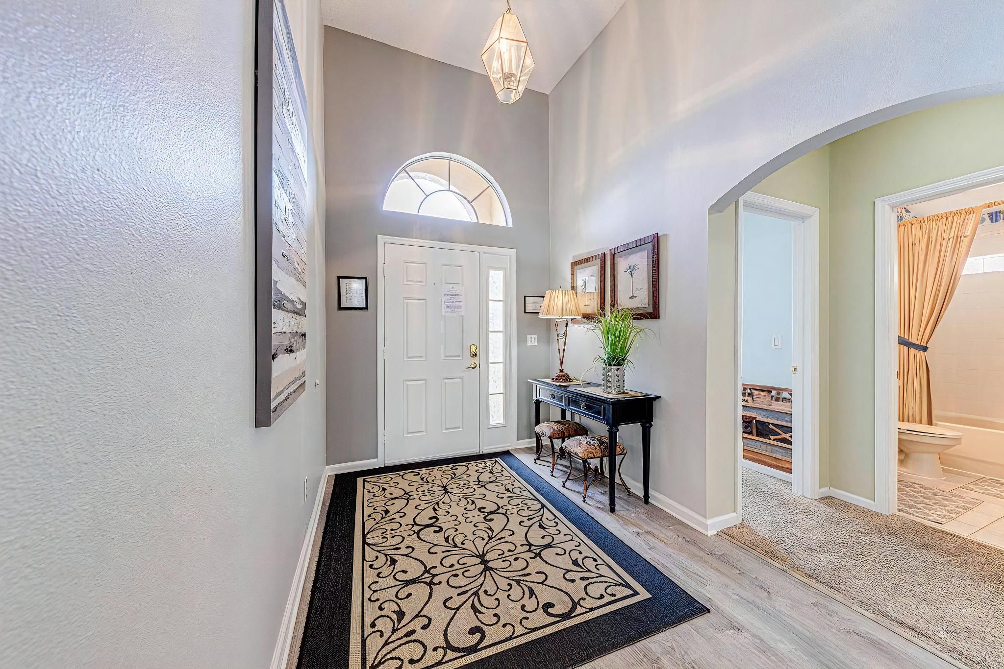A hallway with a white door, a desk with a lamp, framed pictures on the wall, and a patterned rug.
