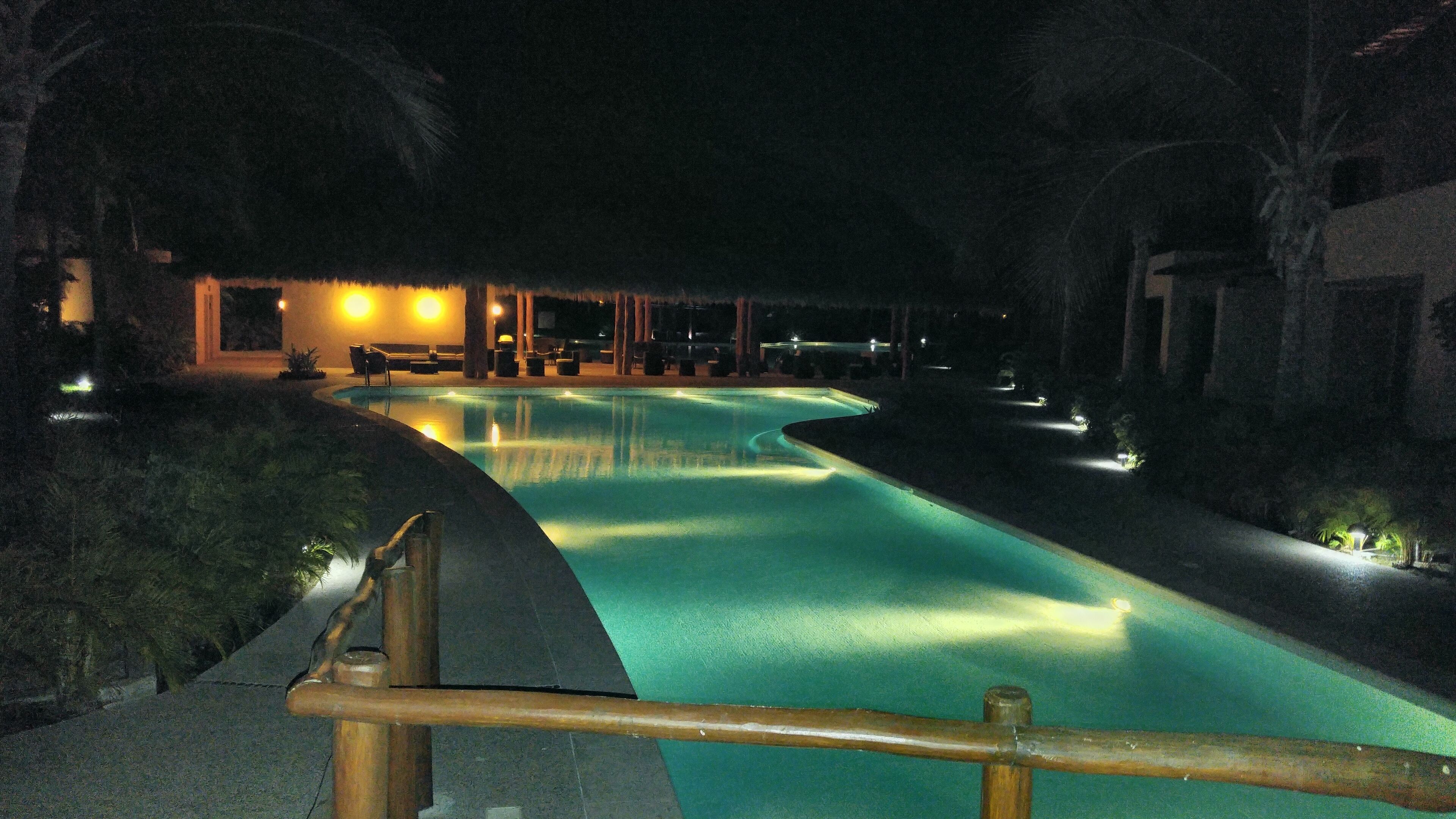 A nighttime swimming pool with a curved design, surrounded by palm trees and a building with outdoor seating.