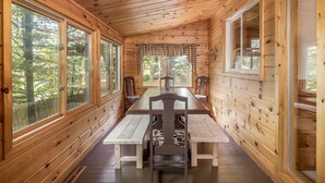 A wooden cabin with a central dining table and chairs, large windows on both sides, and a view of the forest. - Perfect Cabin on Lake Minocqua! (Minocqua)