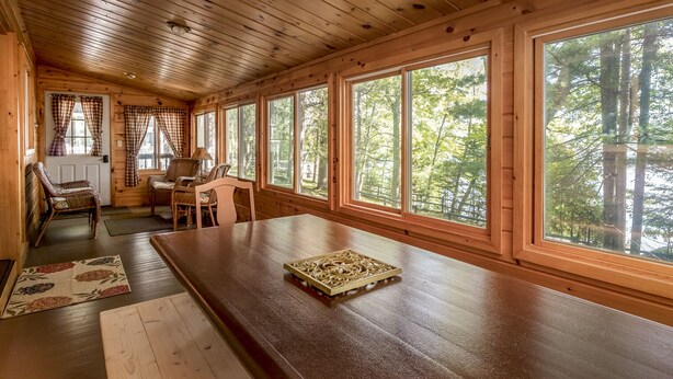 A wooden cabin interior with a long dining table, wicker chairs, and large windows offering a view of the forest. - Perfect Cabin on Lake Minocqua! (Minocqua)
