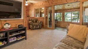 A cozy cabin interior with a television, a bookshelf, a patterned armchair, a sofa, and a sliding glass door leading to an outdoor area. - Perfect Cabin on Lake Minocqua! (Minocqua)