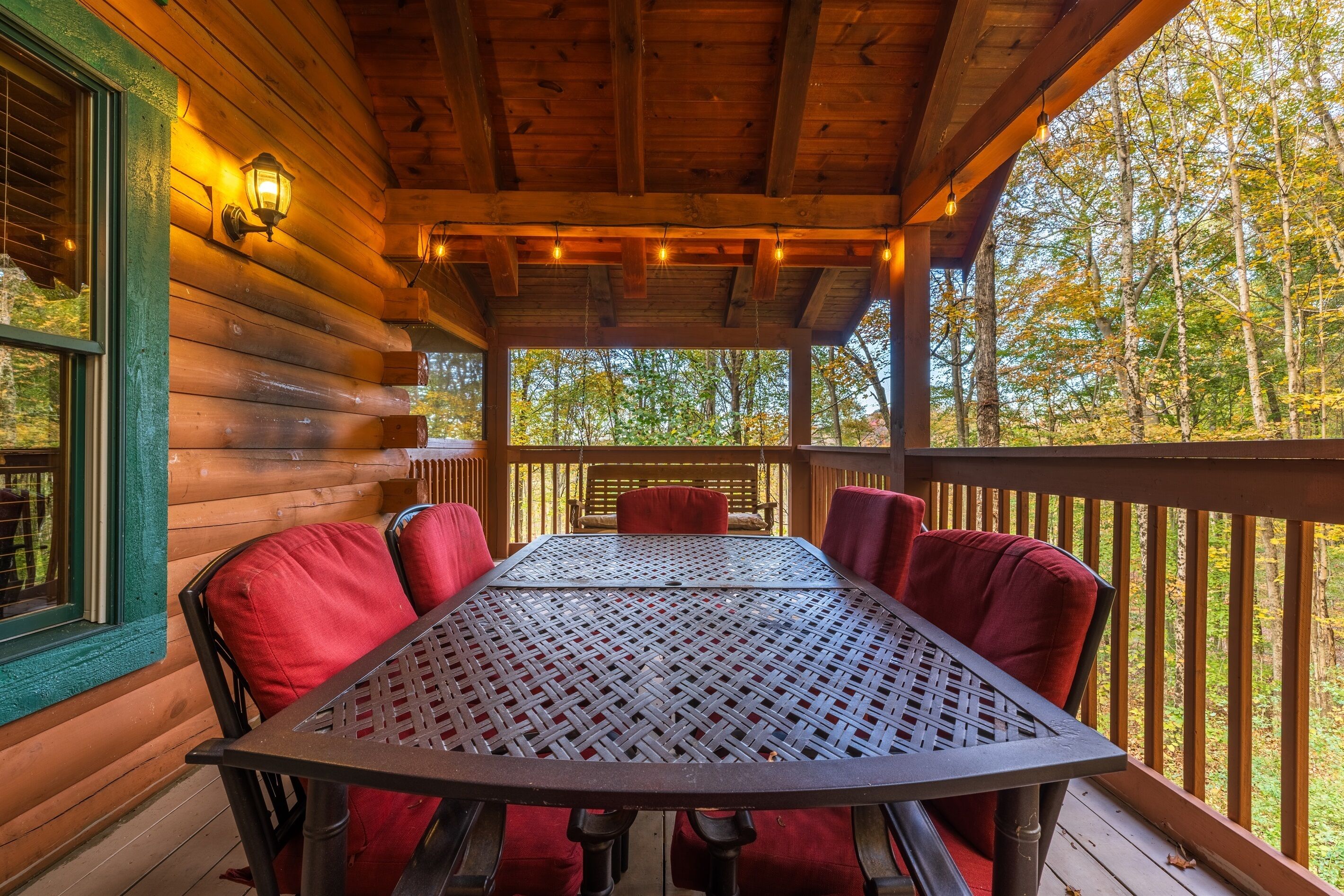 A covered wooden deck with a table and chairs, surrounded by a wooden railing and a view of trees.