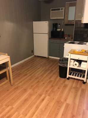 A compact kitchen with a refrigerator, stove, and microwave. There's a wooden table and a trash bin.