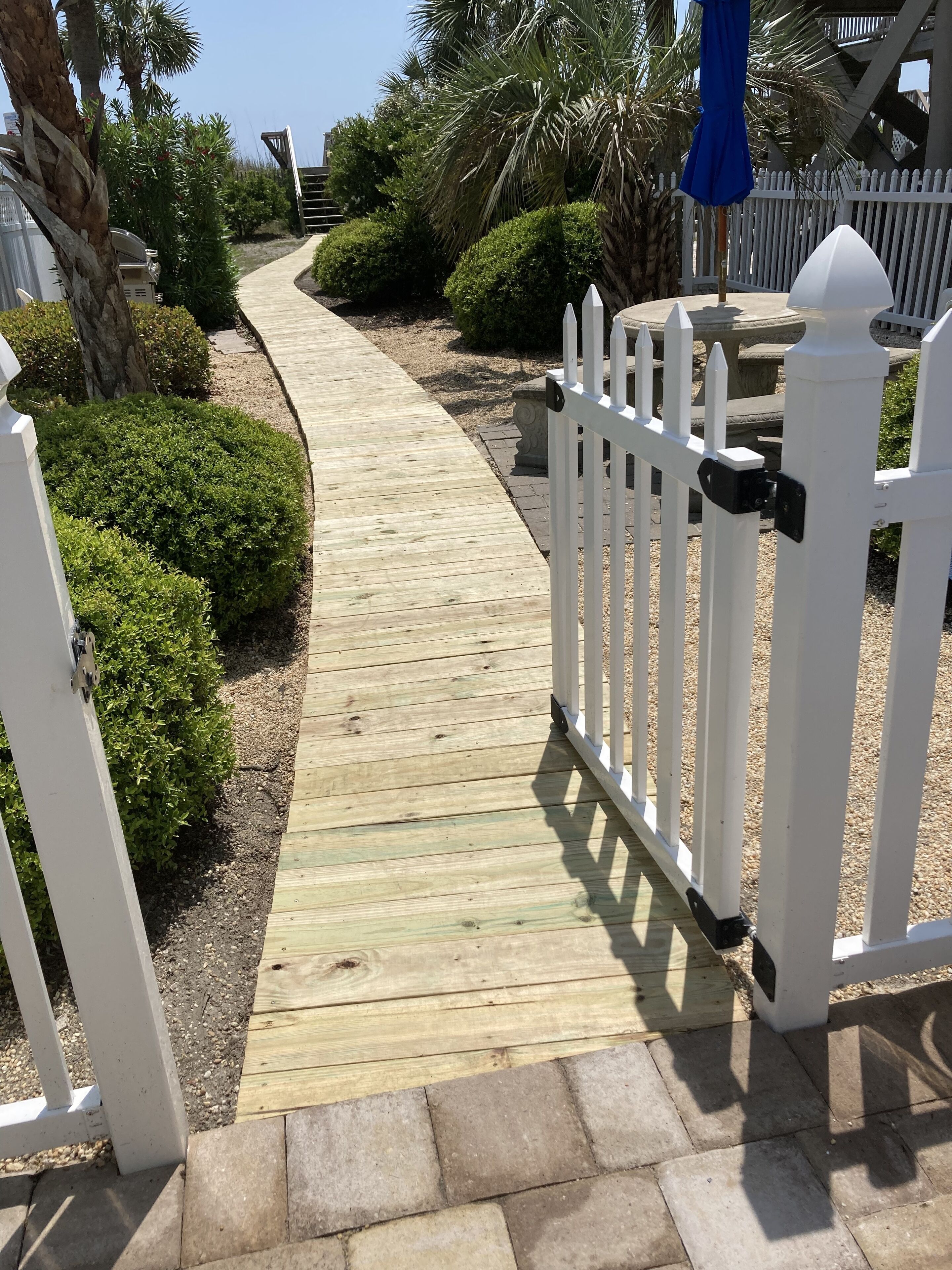 A wooden pathway leading to a gated area with white picket fencing, surrounded by well-maintained bushes and palm trees.