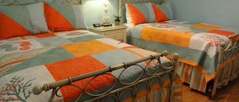 A bedroom with two beds, each with a distinct quilt pattern. There is a nightstand with a lamp and a small plant between the beds. The room has a wooden floor and a light blue wall.