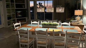 Uma área de refeições com uma mesa de madeira, cadeiras brancas e vista para o pôr do sol através de grandes janelas.