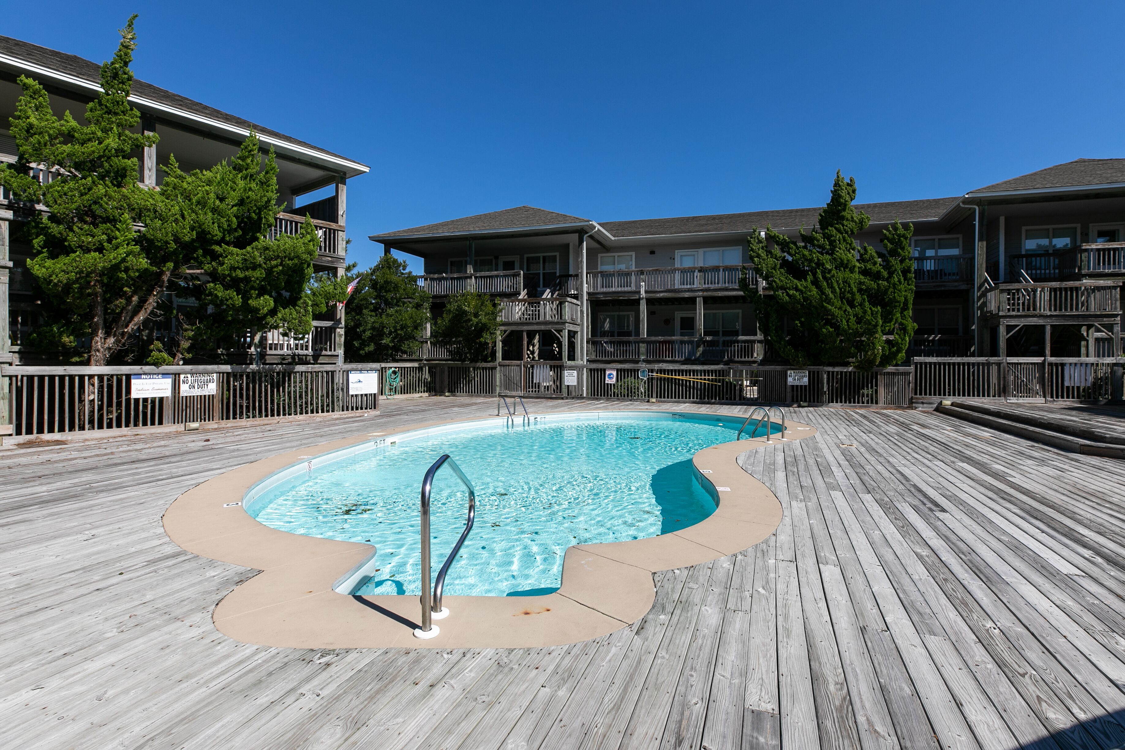 A wooden deck with a swimming pool, surrounded by multi-story buildings with balconies and trees.