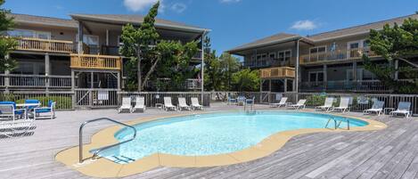 A pool area with a wooden deck, lounge chairs, and a building with balconies.