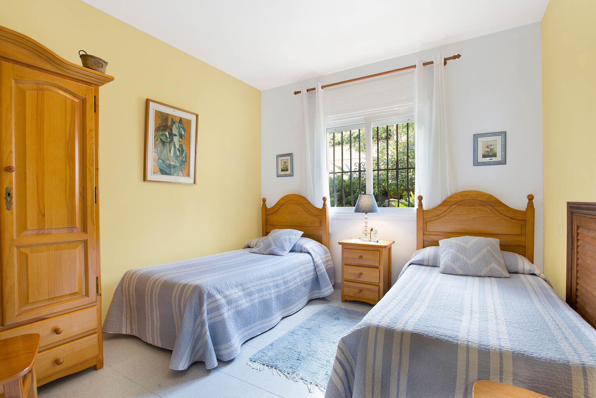 Une chambre à coucher avec deux lits simples, une armoire en bois, une table de chevet avec une lampe et une fenêtre avec des rideaux.