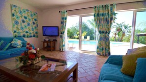 A living room with a sofa, a coffee table with flowers, a TV, and a view of a pool through large windows. - Beautiful Restored Algarve Typical Farmhouse, with Private Pool and Sea Views (Loule)