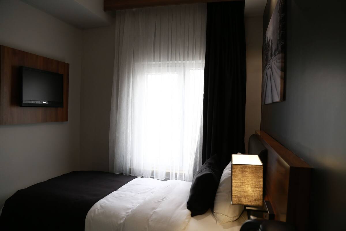 Ein Hotelzimmer mit Bett, einem an der Wand montierten Fernseher, einem Fenster mit transparenten Vorhängen und einem Bild an der Wand.