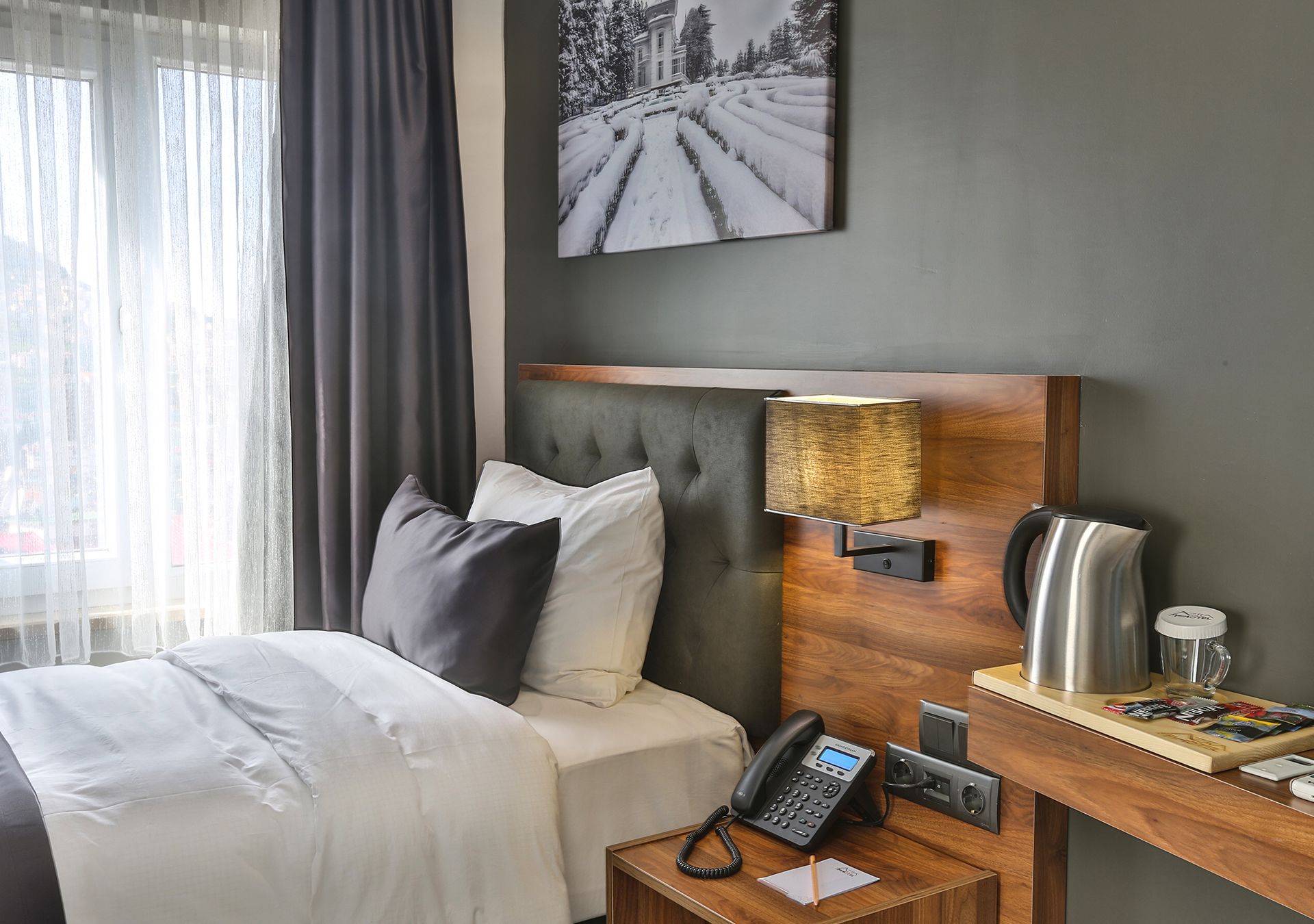 Una habitación de hotel con una cama, una mesita de noche con un teléfono, una lámpara, una cafetera y un cuadro de un paisaje nevado.