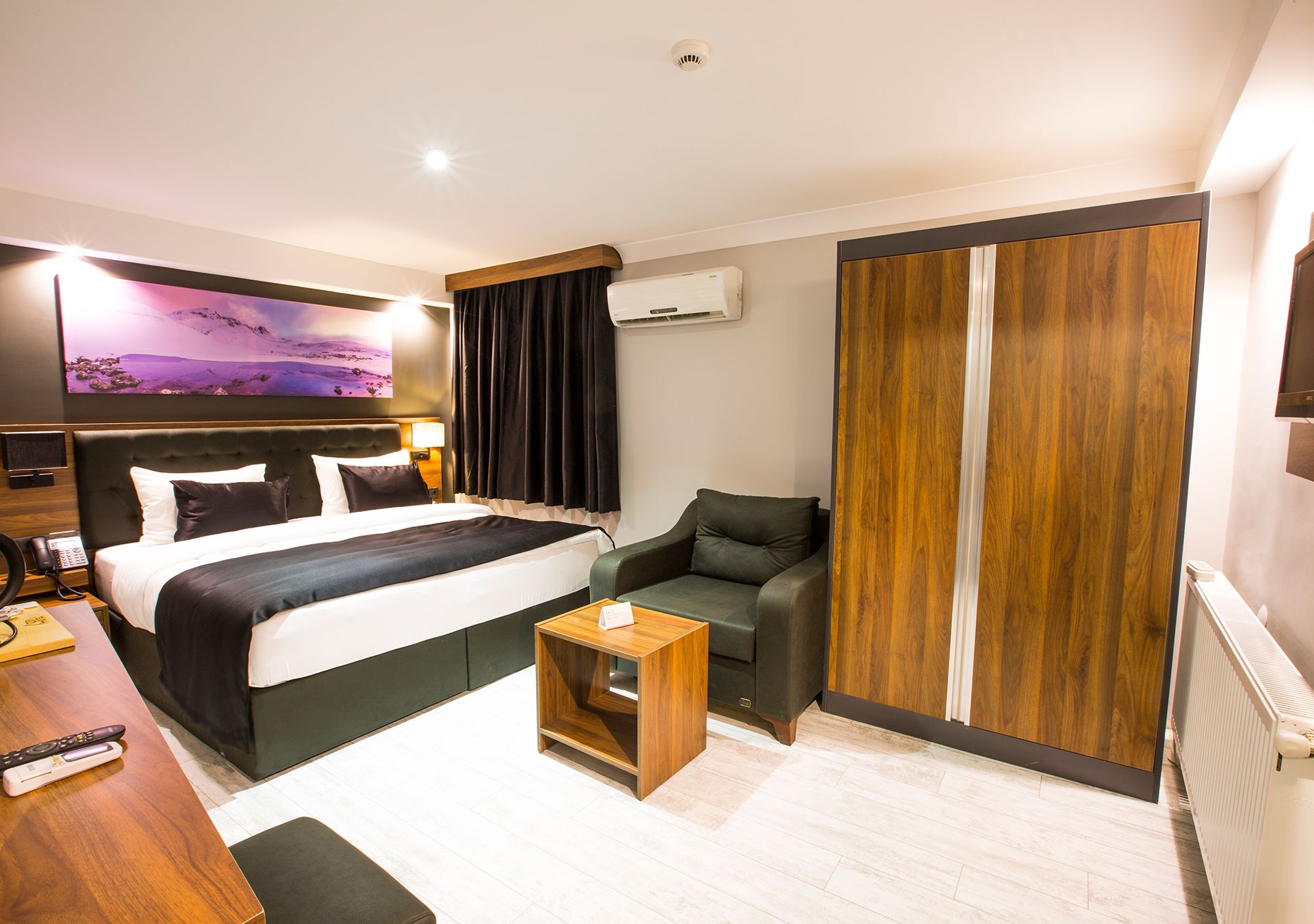 A hotel room with a large bed, a wooden side table, a leather armchair, a large window with curtains, and a mounted air conditioner.