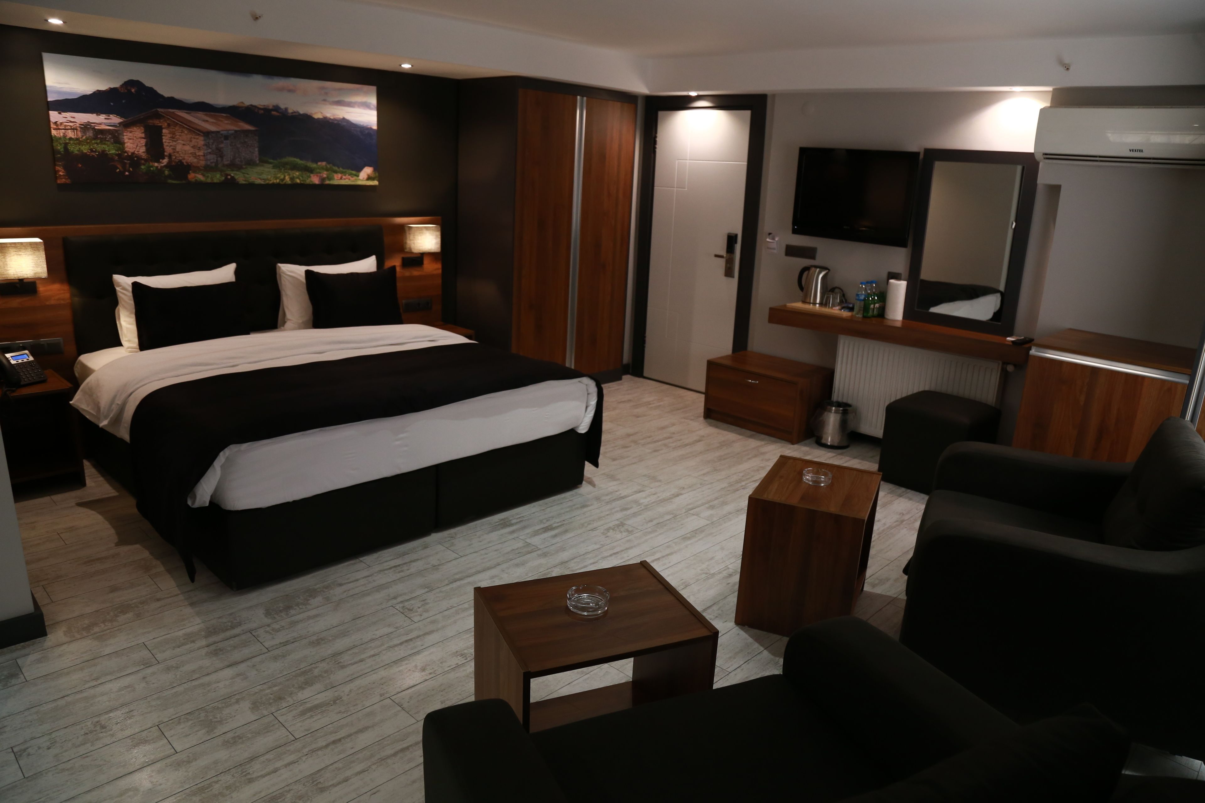 Habitación de hotel moderna con una cama grande, una mesita de noche de madera, una pequeña mesa, una silla, un televisor y un espejo.