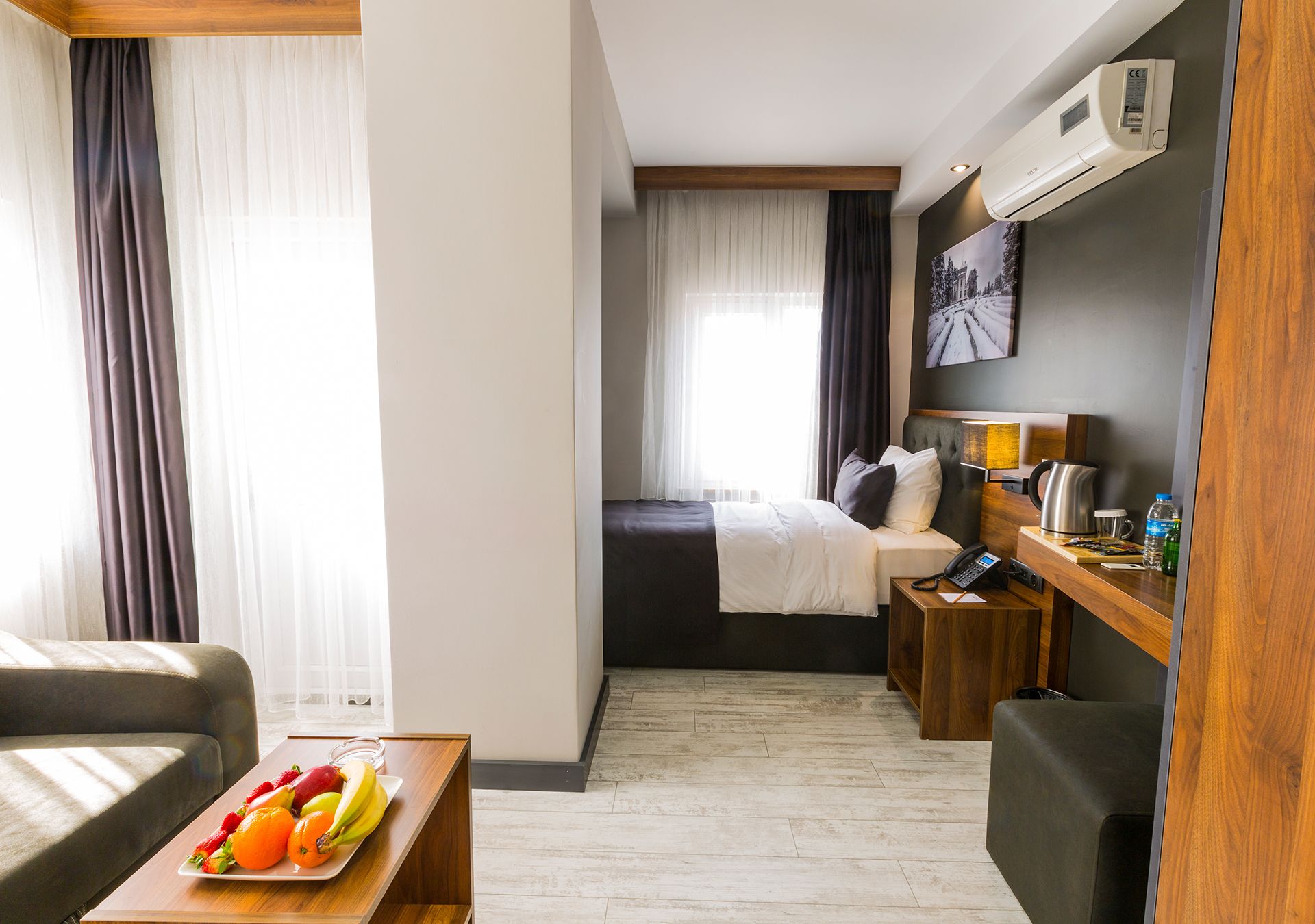 Quarto de hotel com cama, escrivaninha com telefone, mesinha com frutas e ar-condicionado fixado na parede.