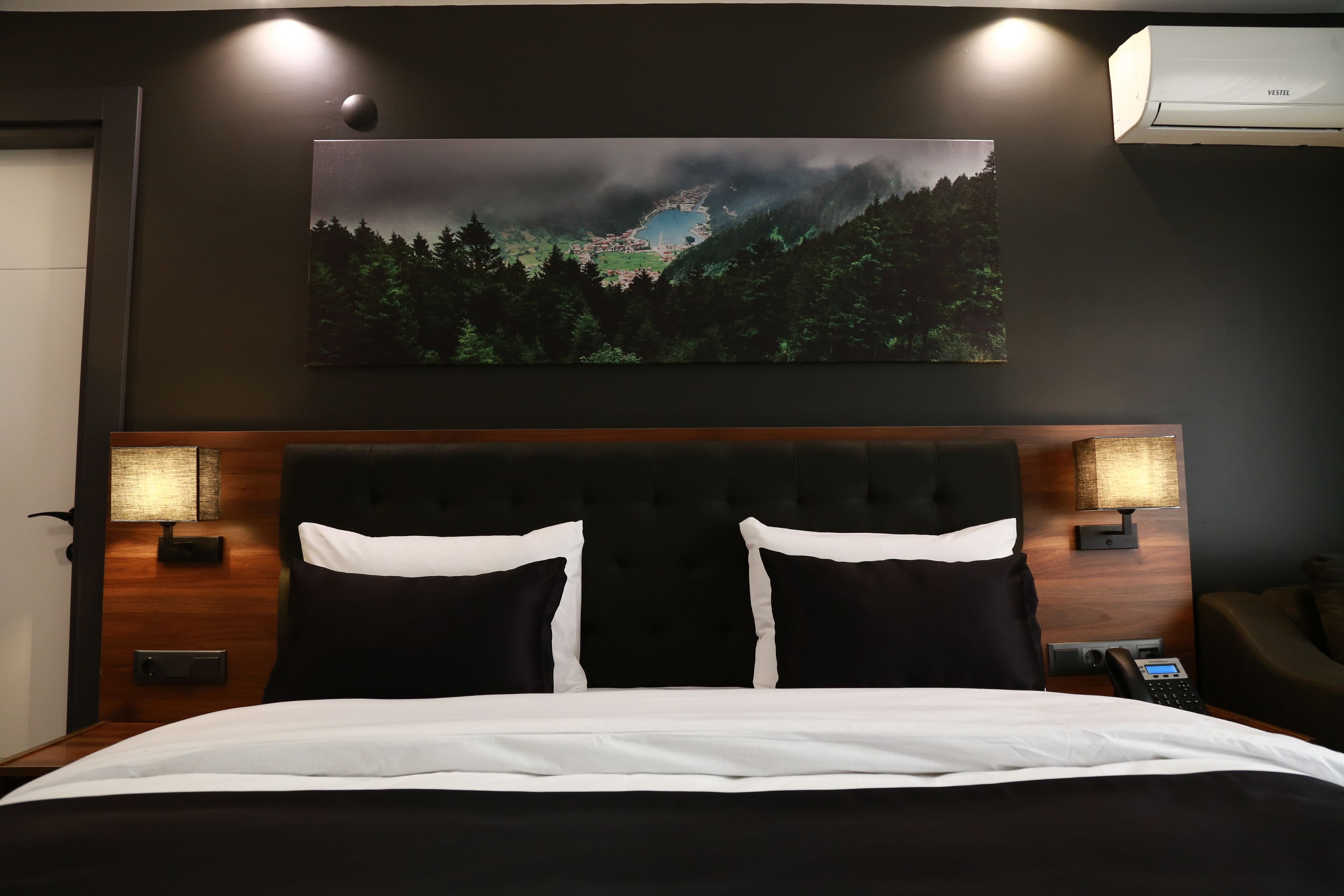 Une chambre d’hôtel avec un grand lit, deux tables de chevet, une petite table, un climatiseur fixé au mur et un tableau représentant un paysage forestier.