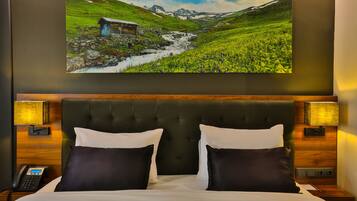 Een hotelkamer met een groot bed, twee bedlampen, een houten hoofdeinde en een lijst met een berglandschap aan de muur.