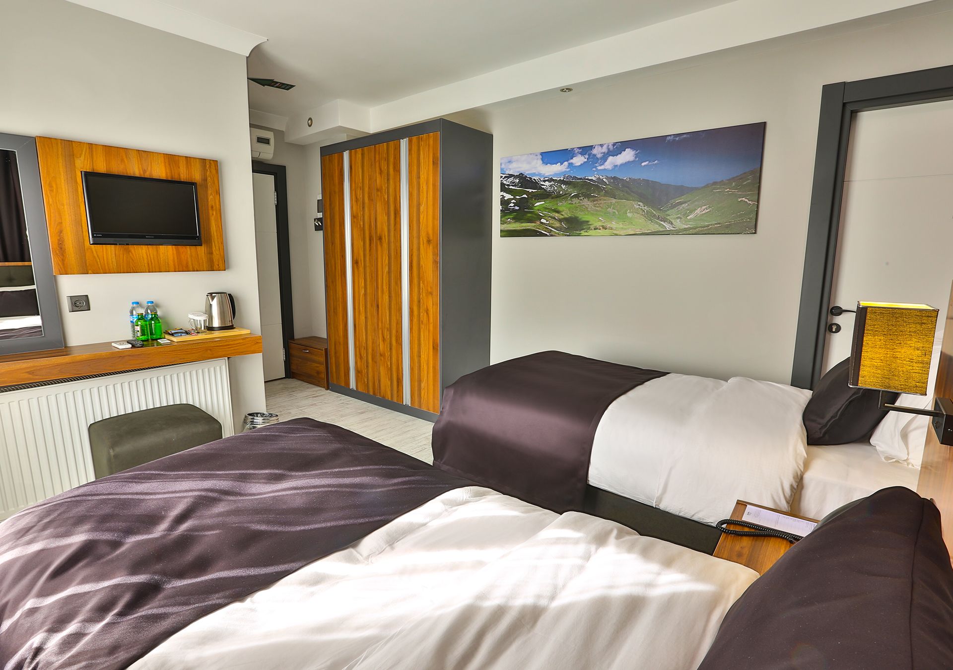 A hotel room with two beds, a desk with a flat-screen TV, a wooden wardrobe, and a mountain landscape painting on the wall.