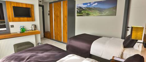 A hotel room with two beds, a desk with a flat-screen TV, a wooden wardrobe, and a mountain landscape painting on the wall.