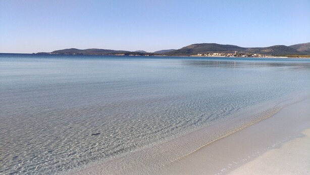 A beach with clear shallow waters, a sandy shore, and a distant town with buildings and hills. - PENTHOUSE MARIS ALGHERO VIALE SARDINIA 104 LUXURY PENTHOUSE NEAR THE BEACH (Alghero)