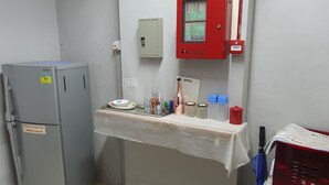 A small kitchenette with a refrigerator, a fire alarm panel, and a table with various items. - Century Service World - Hostel (Singapore)