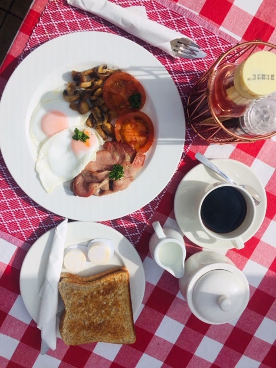 A breakfast setting with eggs, mushrooms, and toast on a plate, a cup of coffee, and a bottle of milk on a checkered tablecloth.