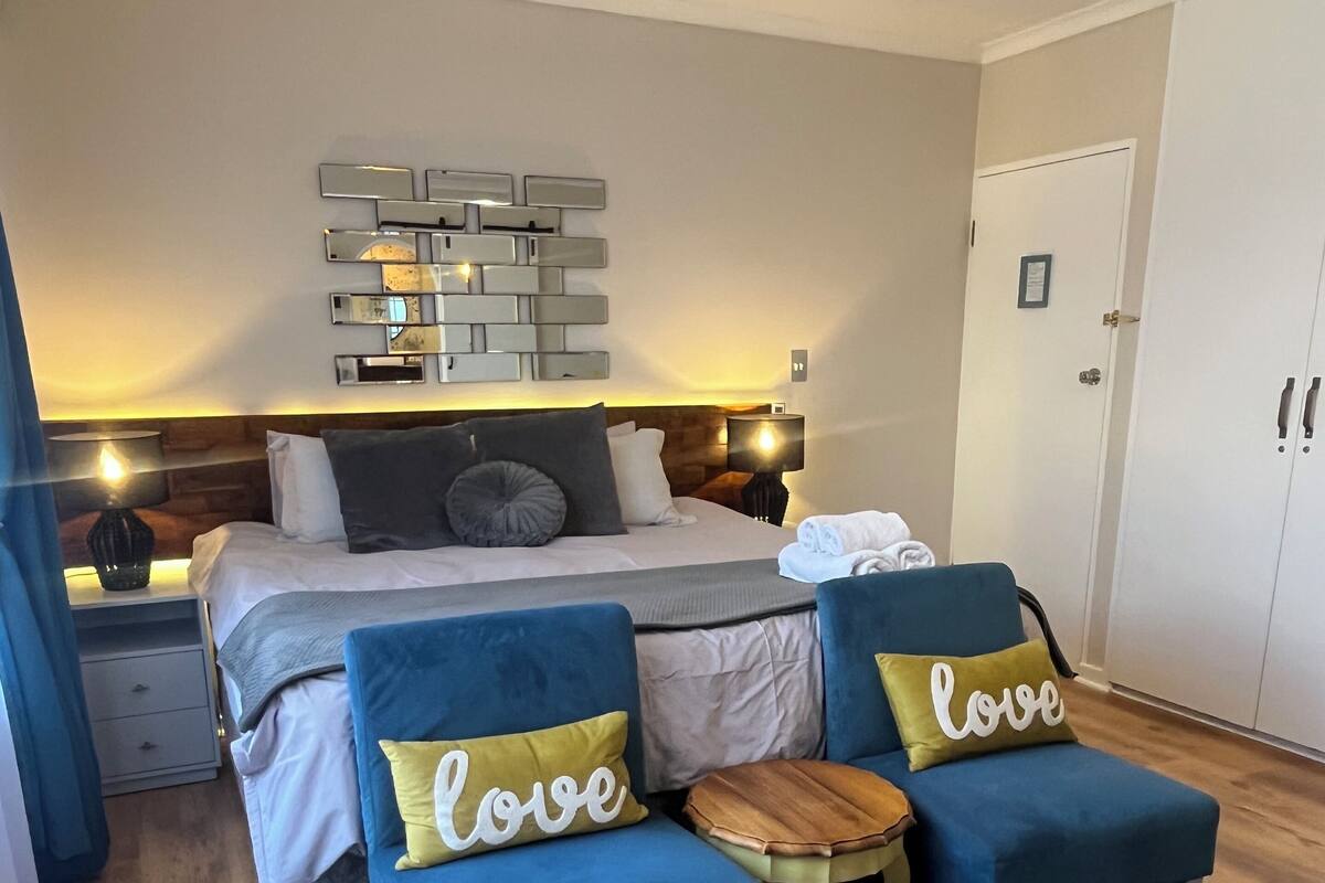 A bedroom with a bed, two blue armchairs, a wooden side table, a blue rug, a ceiling fan, and a wall-mounted light fixture.