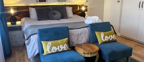 A bedroom with a bed, two blue armchairs, a wooden side table, a blue rug, a ceiling fan, and a wall-mounted light fixture.