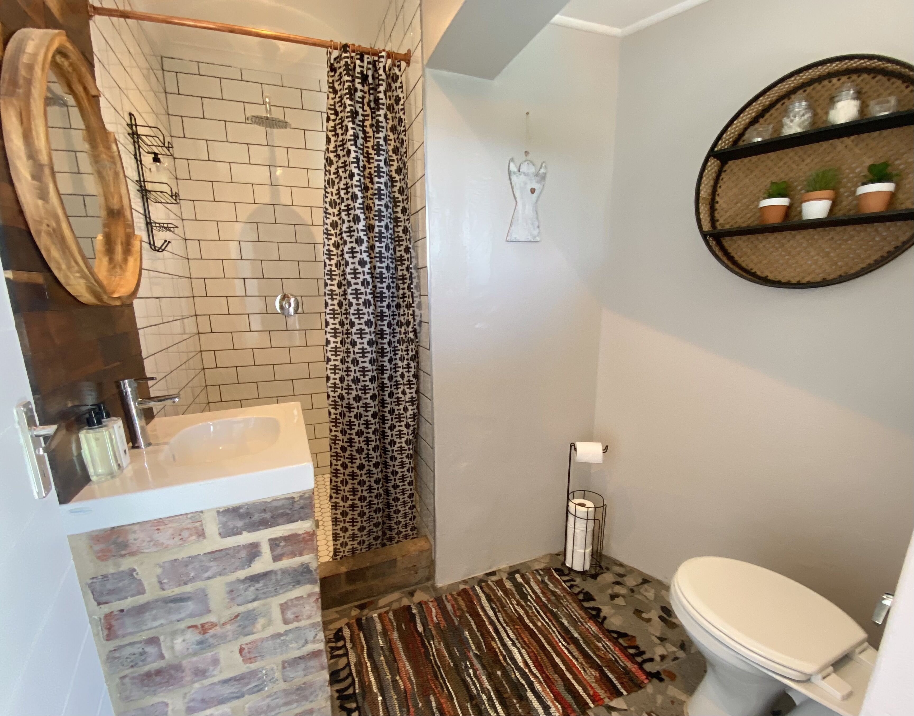 Ein Badezimmer mit Dusche, Toilette, Waschbecken, Spiegel, einem gemusterten Duschvorhang, einem Wanddekor und einem Regal mit kleinen Pflanzen.