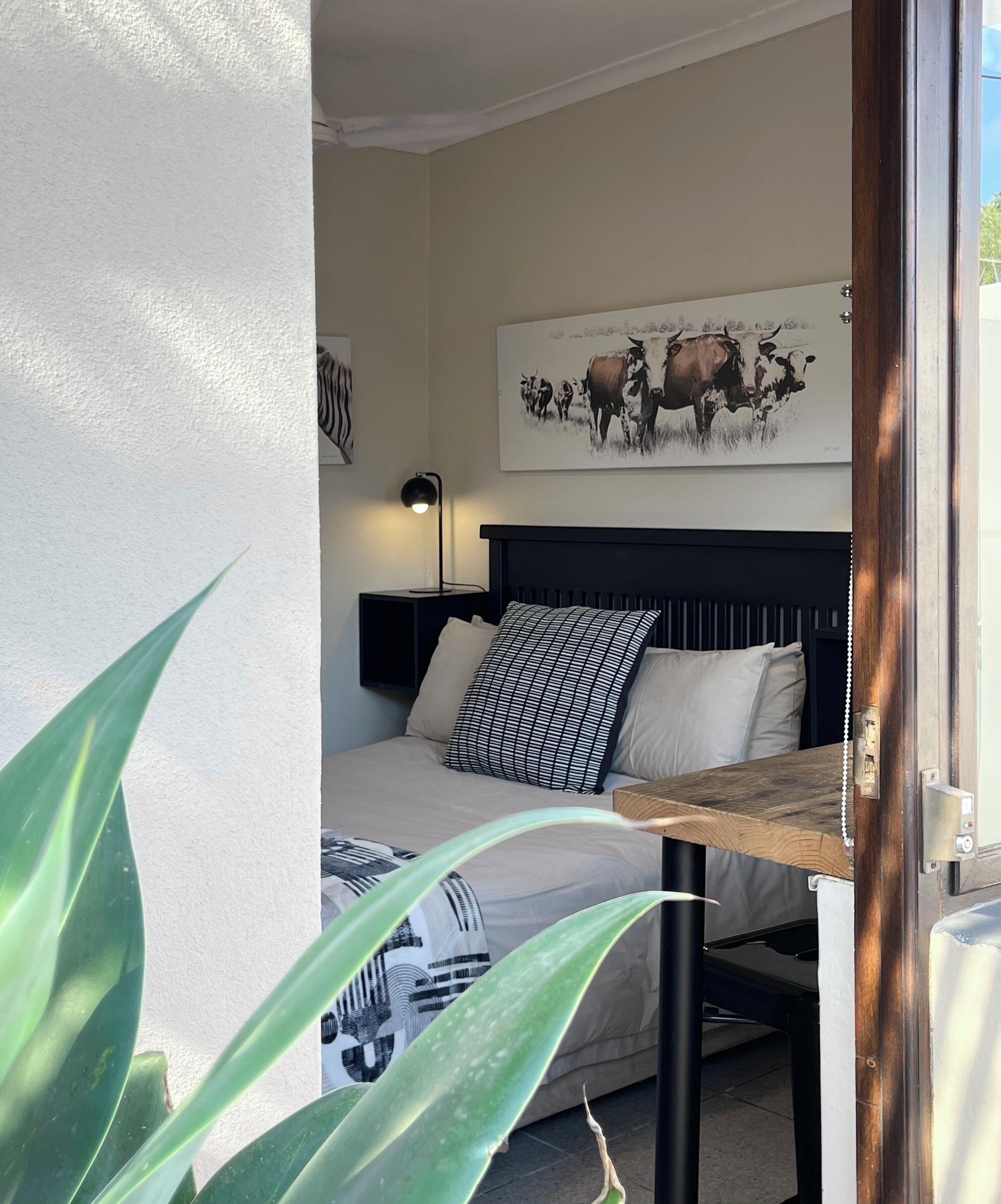 A bedroom with a bed, a nightstand, a lamp, a painting, and a plant.