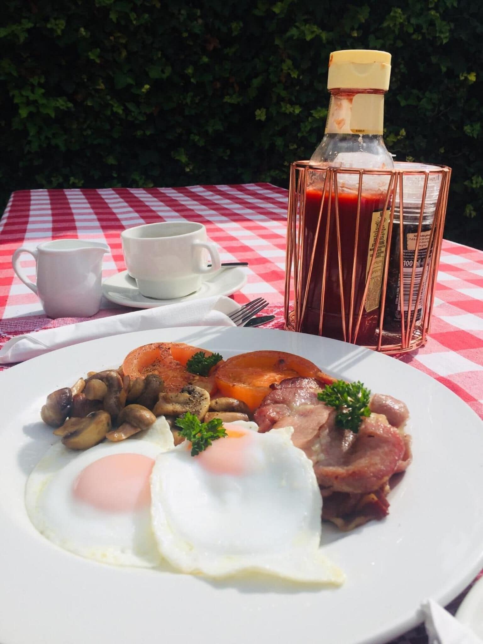 Un plato de desayuno con huevos, jamón, champiñones y tomates, una jarra de jugo y una taza blanca sobre un mantel a cuadros.