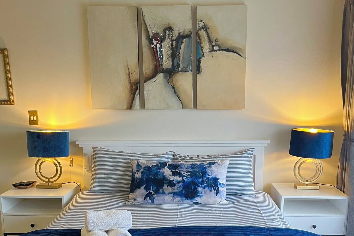 A neatly made bed with striped bedding and a floral pillow, flanked by two bedside tables with lamps, a wall-mounted artwork, and a visible power outlet.