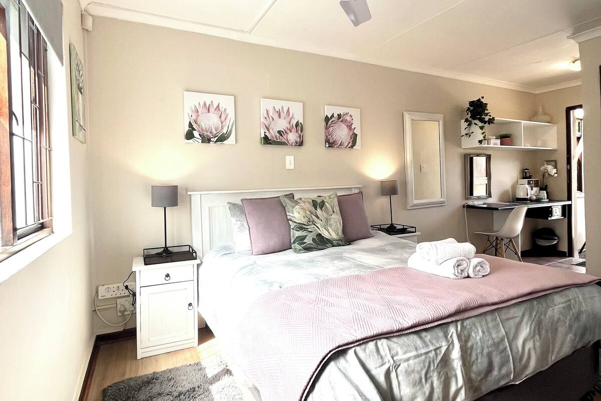 A bedroom with a bed, bedside tables, a nightstand, a desk, a chair, a ceiling fan, and a window.