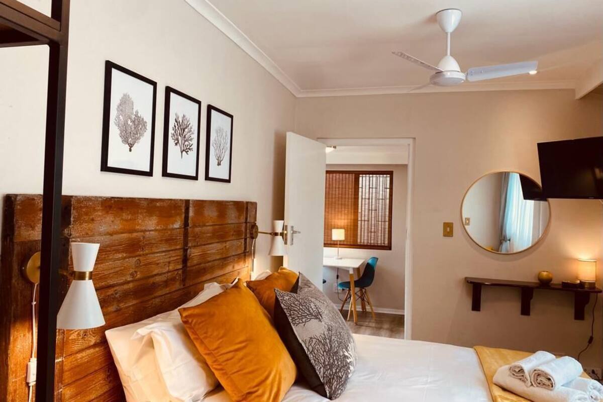 A hotel room with a wooden headboard, a bed with white and orange bedding, a desk with a chair, a mirror, and framed botanical prints on the wall.