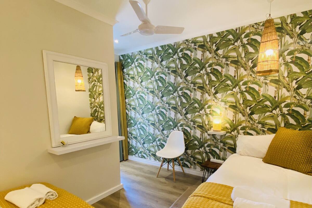A hotel room with two beds, a ceiling fan, a large mirror, and a wall with a leafy pattern.