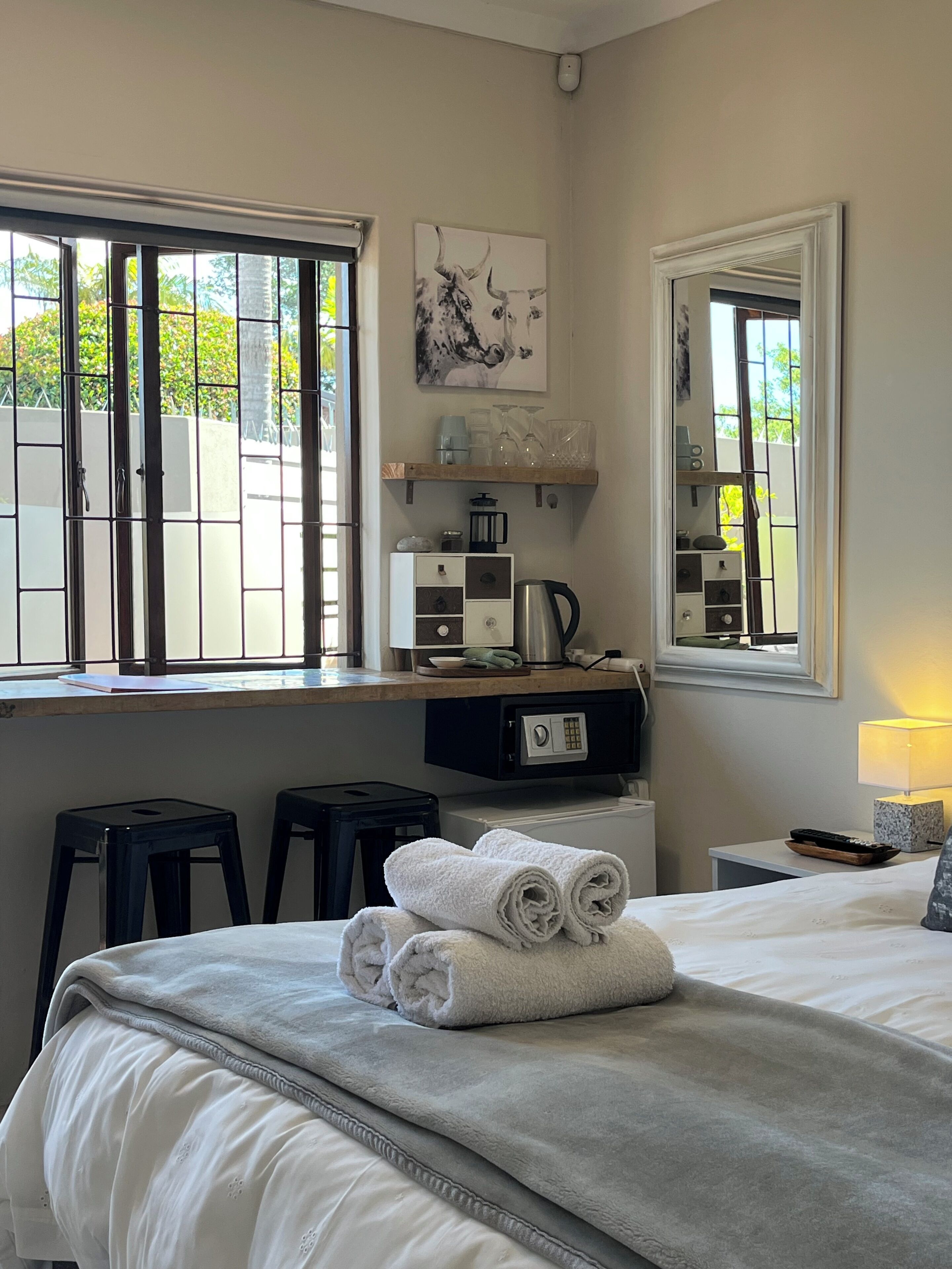A hotel room with a bed, a window with bars, a coffee maker, a mirror, and a small table with a lamp.