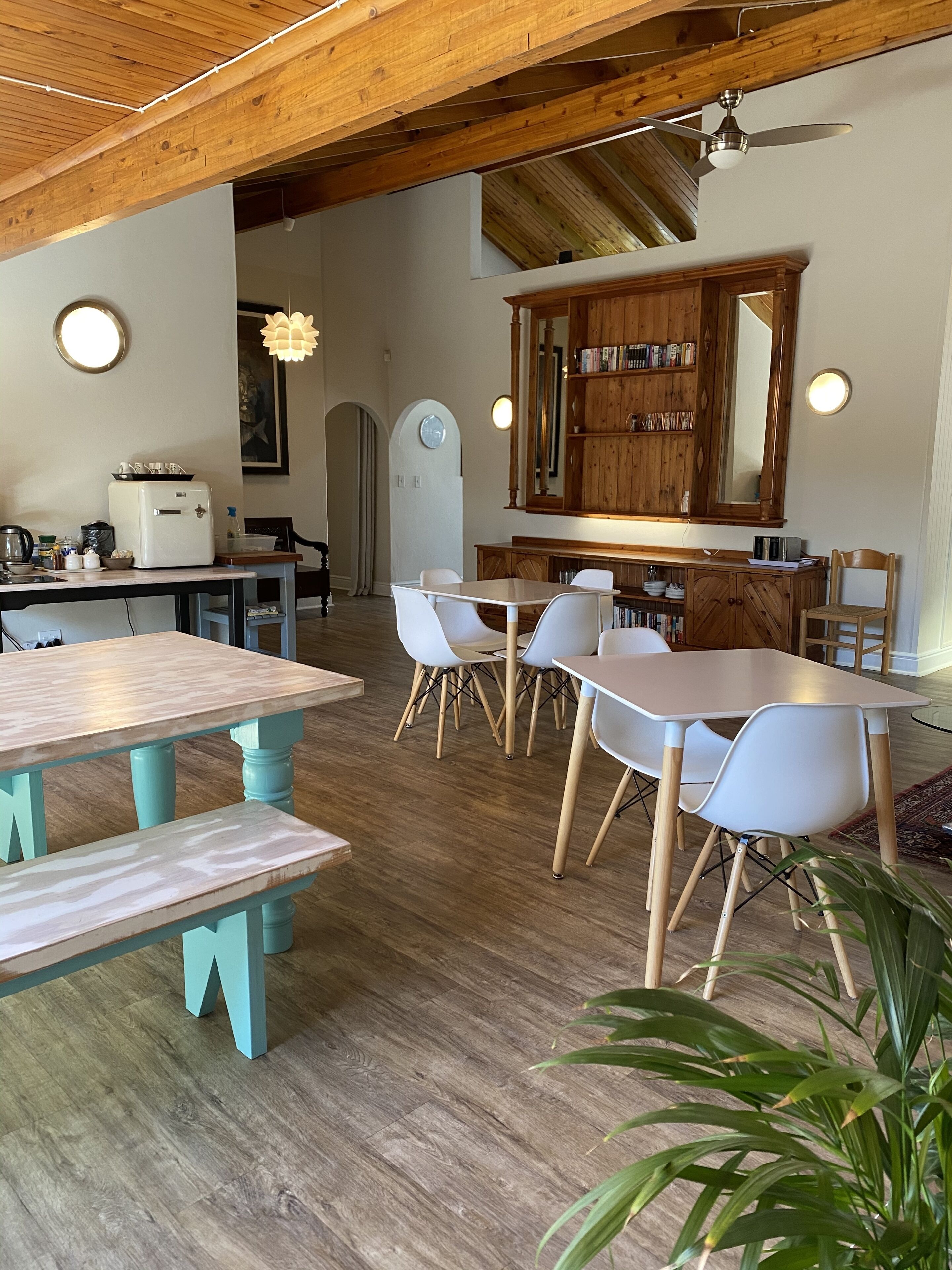 A dining area with wooden tables and chairs, a kitchen area with a refrigerator and a sink, and a large mirror above a wooden cabinet.