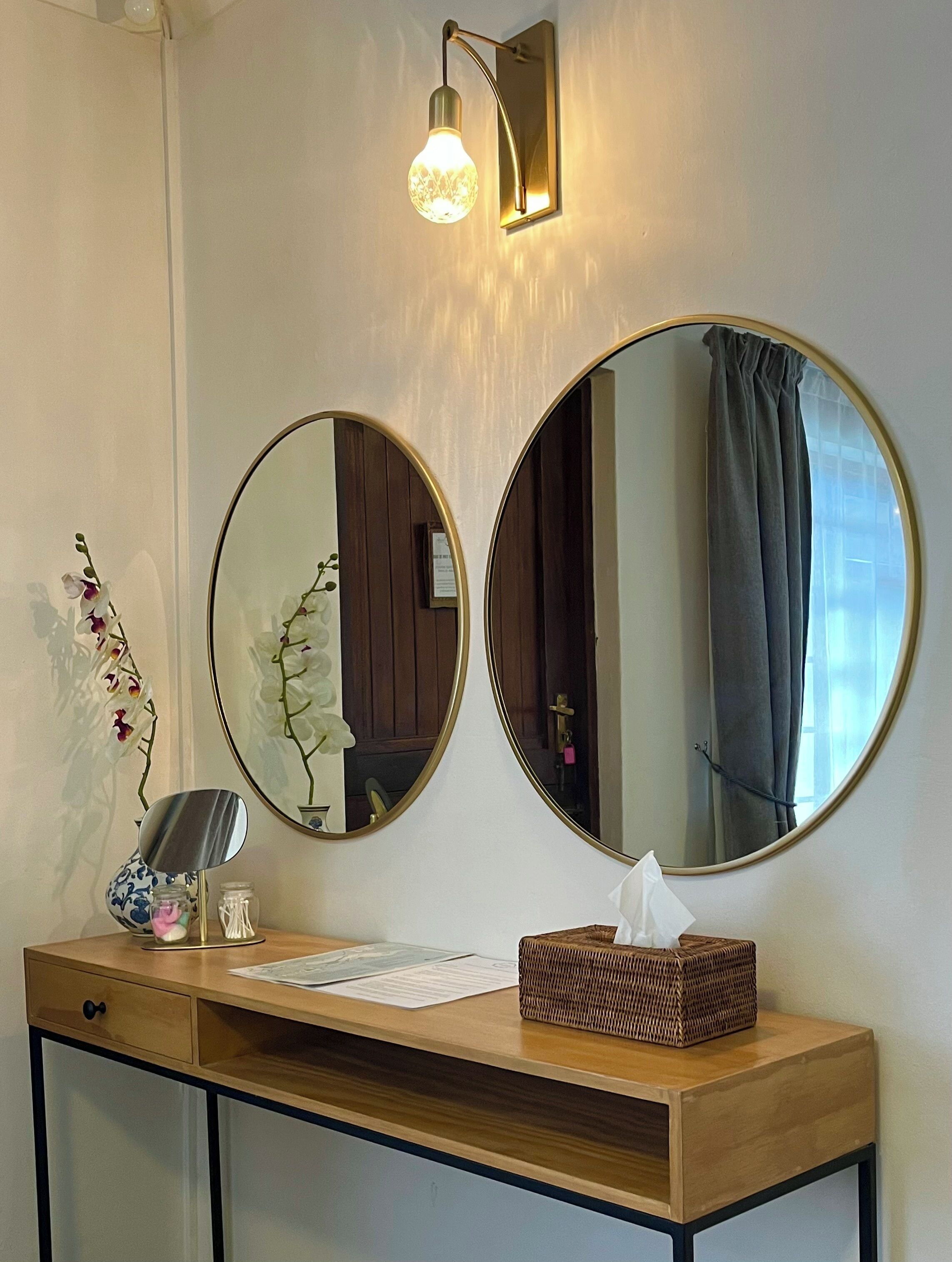 A wooden console table with two round mirrors, a small vase with flowers, a tissue box, and a wall-mounted light fixture.