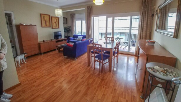 A spacious living area with a wooden floor, a blue sofa, a dining table with chairs, a TV, and a balcony with a view. - Family Apartment Just Steps from the Sea in Palamós 🏖️☀️ (Palamós)