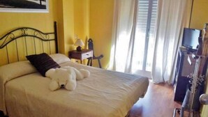 A bed with a white bedspread and a white teddy bear, a nightstand with a lamp, a TV on a stand, and a window with curtains. - Family Apartment Just Steps from the Sea in Palamós 🏖️☀️ (Palamós)