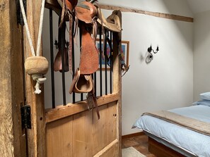 A rustic wooden stable with leather tack, a bed with a blue blanket, and a framed picture on the wall.