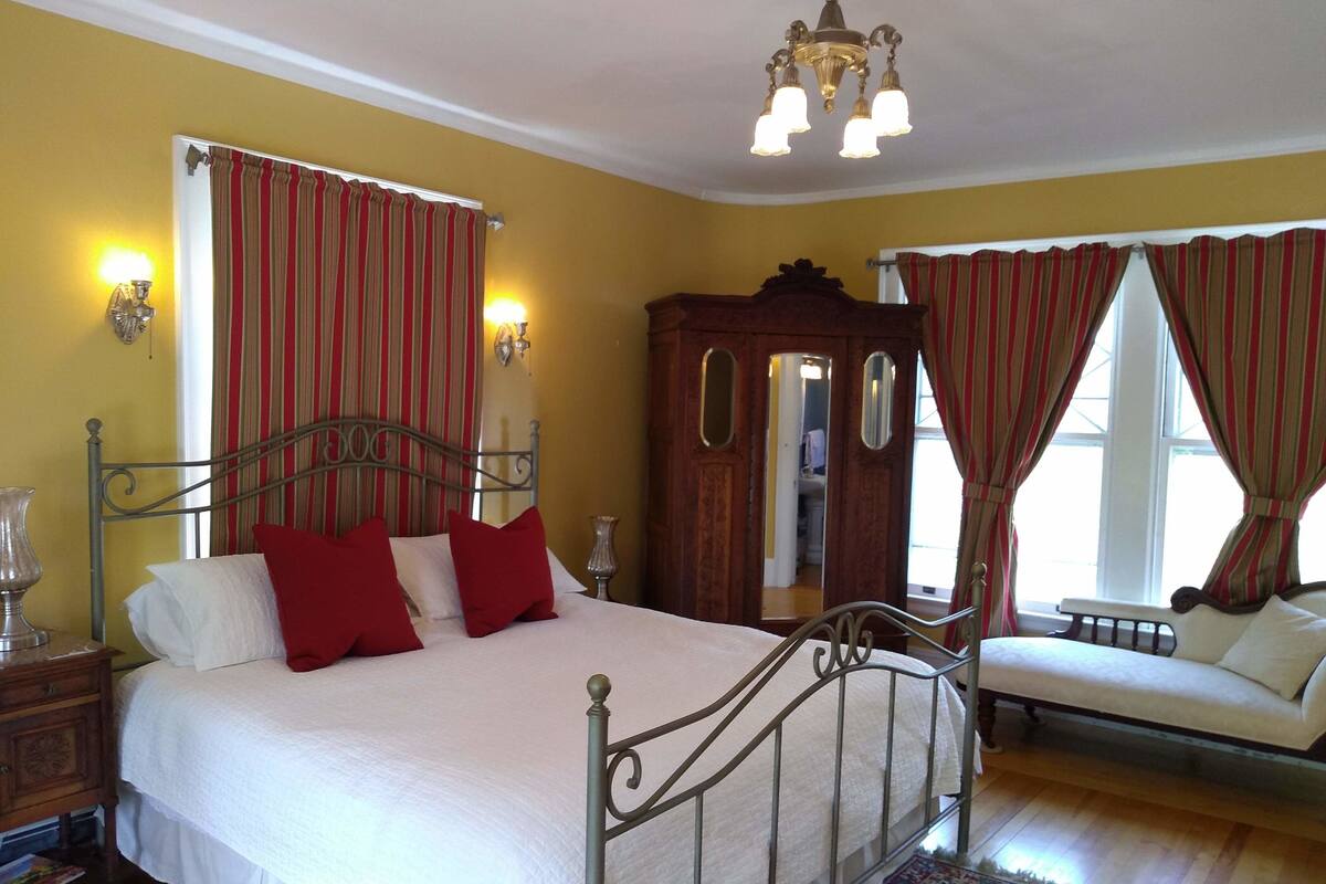 A bedroom with a metal bed frame, a wooden nightstand, a bedside table, a mirror, and a window with red curtains.