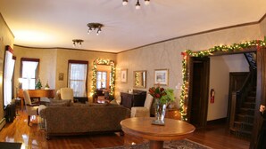 Un salón acogedor con suelo de madera, una mesa redonda de centro y una chimenea. Hay dos sofás y una mesa de comedor con sillas. La habitación está decorada con luces navideñas y una guirnalda.
