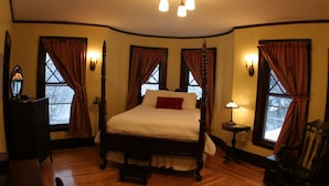 Un dormitorio tradicional con cama con dosel, suelo de madera y vistas al exterior a través de ventanas con cortinas.