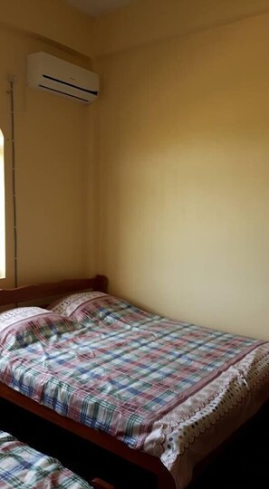 A single bed with a plaid bedspread and a white lace border, an air conditioning unit mounted on the wall, and a window on the left side.