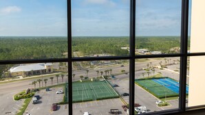 A view through a window of a parking lot with a tennis court, a road, and a forested area in the background. - Wonderful Location! Clean Unit with Gorgeous Gulf Front Views! Family Friendly! (Orange Beach)