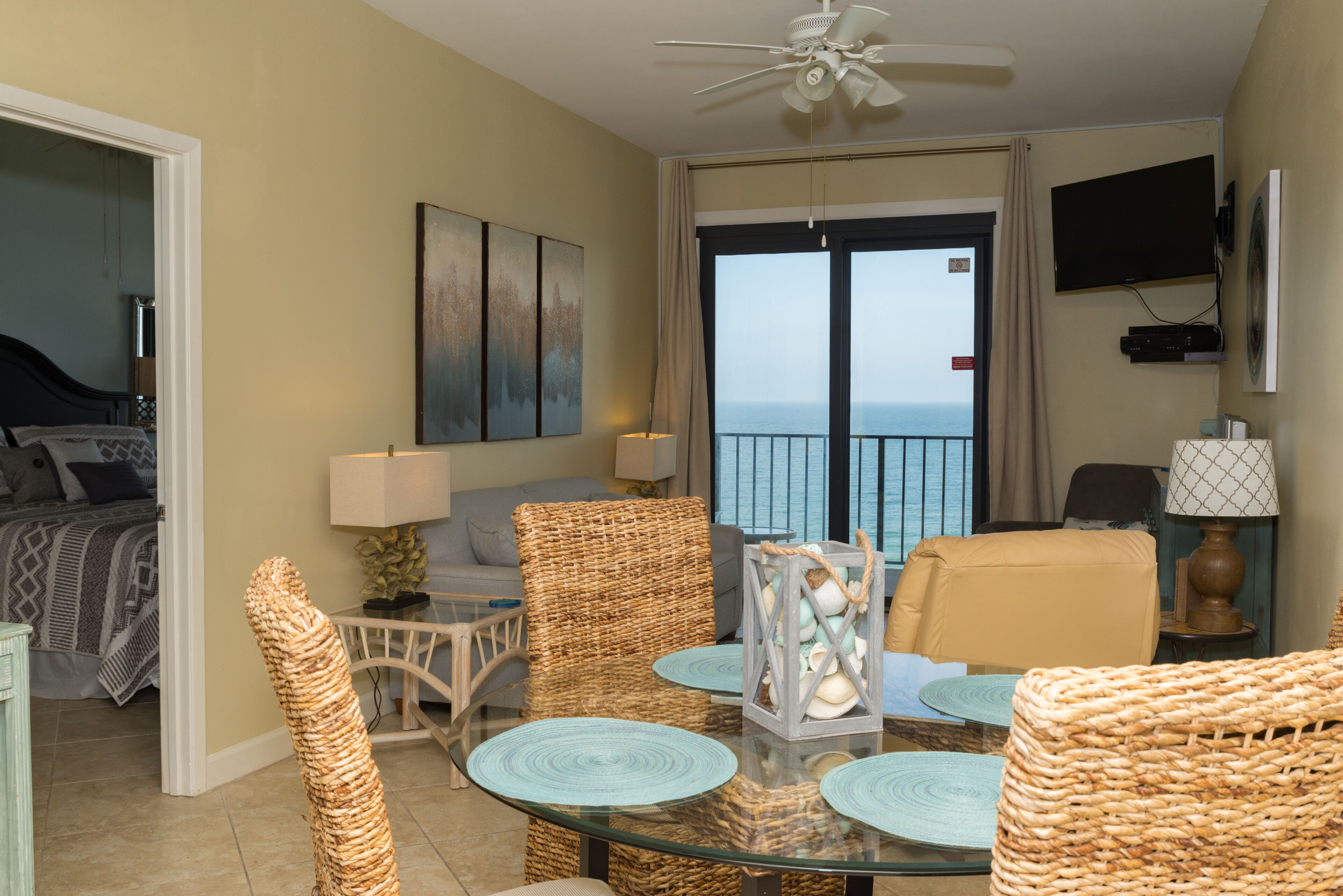 A room with a glass table, wicker chairs, a bed with patterned bedding, a TV mounted on the wall, and a view of the sea through a large window.