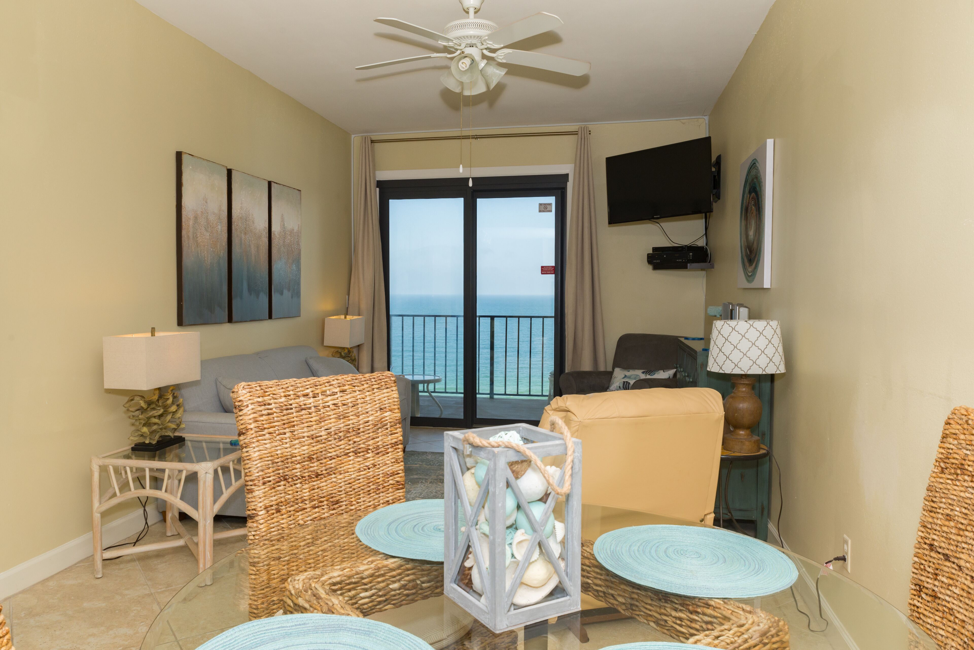 A room with wicker furniture, a glass table, a ceiling fan, a TV, and a view through a glass door to the outside.