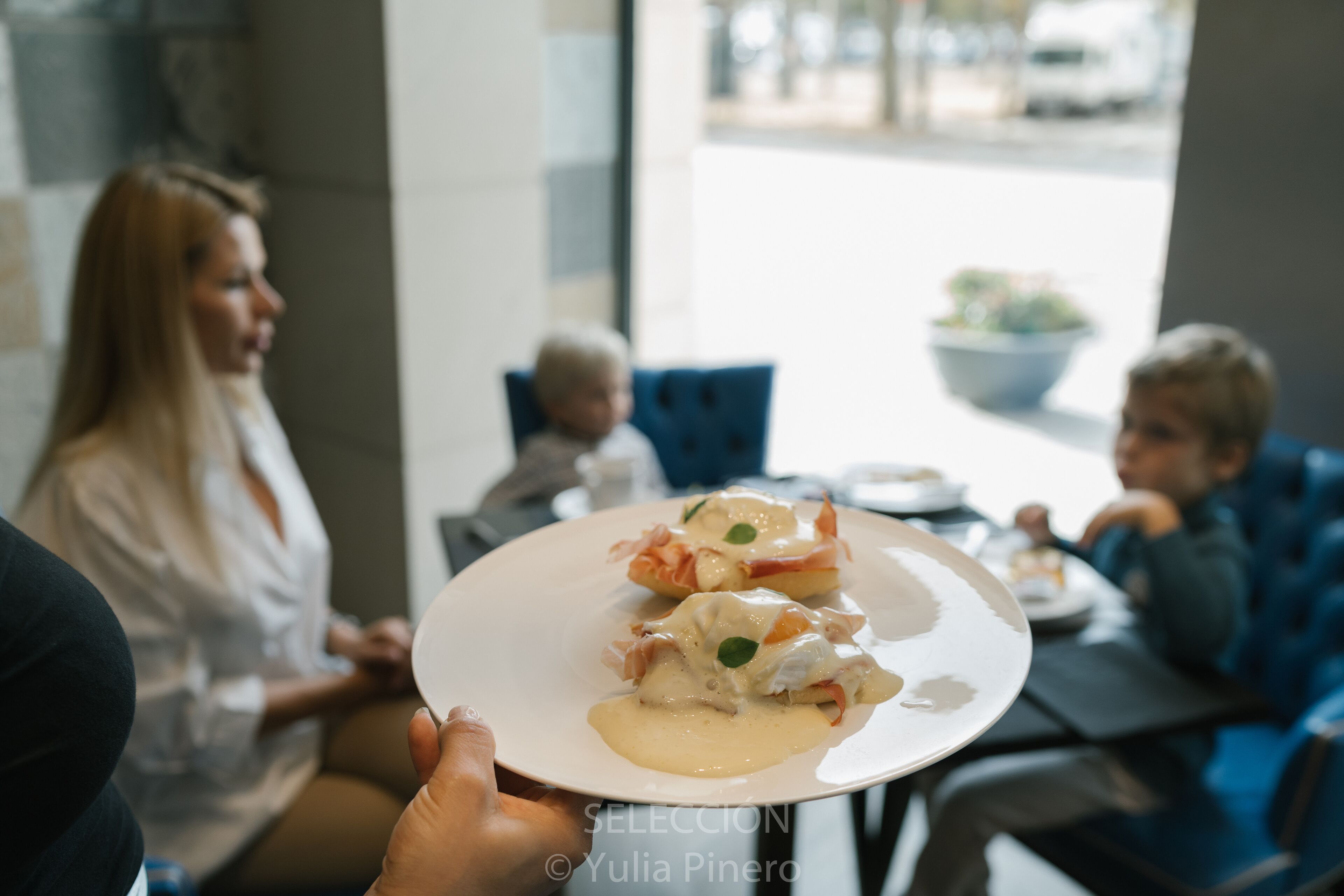 Una mesa de comedor con un plato de comida, una persona sosteniendo un plato y un niño al fondo.